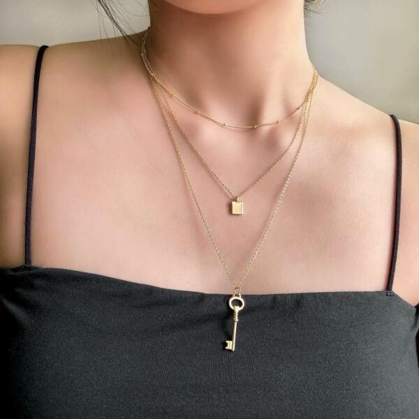 Model wearing layered gold necklaces, including a choker, square pendant, and key charm, styled for a trendy, modern look.